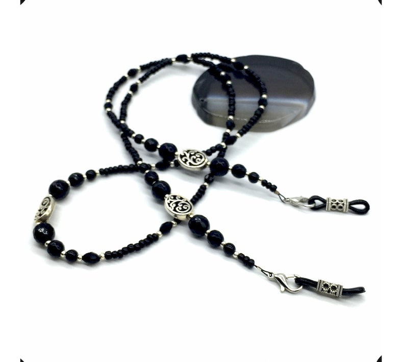 May include: Black beaded eyeglass chain with silver accents. The chain features small black beads and larger black beads with silver filigree designs. The chain has two silver clasps for attaching to eyeglasses.