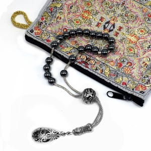Puede incluir: Un rosario de cuentas gris oscuro con una borla decorativa plateada y una cuenta plateada. Las cuentas est&aacute;n sobre una bolsa gris con un estampado floral y una cadena dorada. La bolsa tiene una cremallera negra.