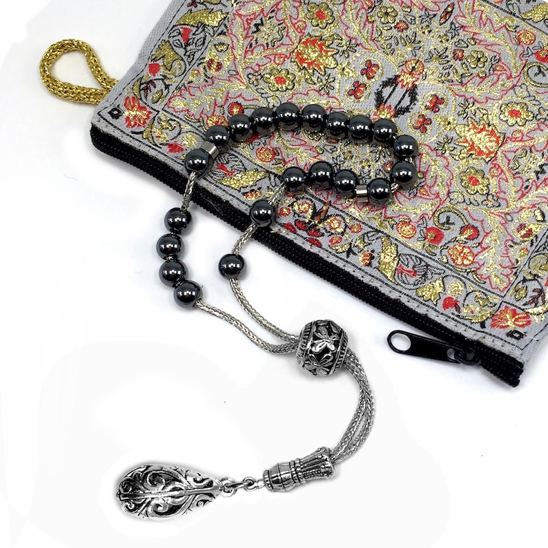 Puede incluir: Un rosario de cuentas gris oscuro con una borla decorativa plateada y una cuenta plateada. Las cuentas est&aacute;n sobre una bolsa gris con un estampado floral y una cadena dorada. La bolsa tiene una cremallera negra.