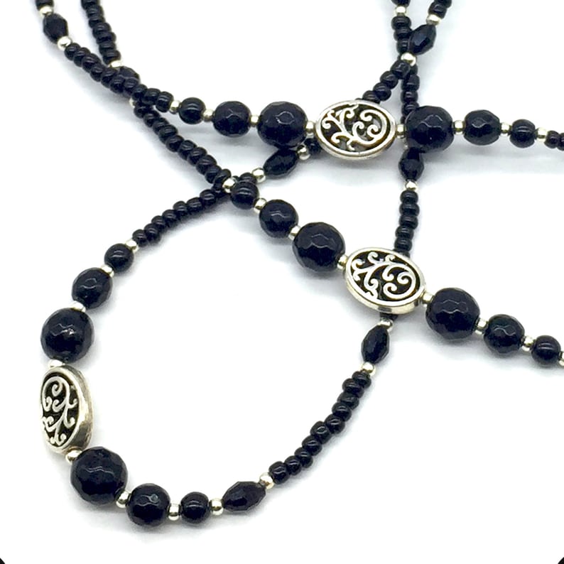 May include: A black beaded necklace with silver oval charms featuring a floral design. The necklace is made of two strands of beads, one with larger beads and one with smaller beads.