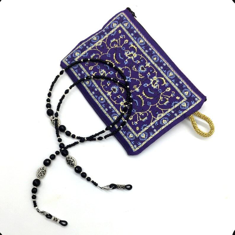 May include: Black beaded eyeglass chain with silver accents and a purple patterned pouch. The chain has a double strand of black beads and silver oval charms.