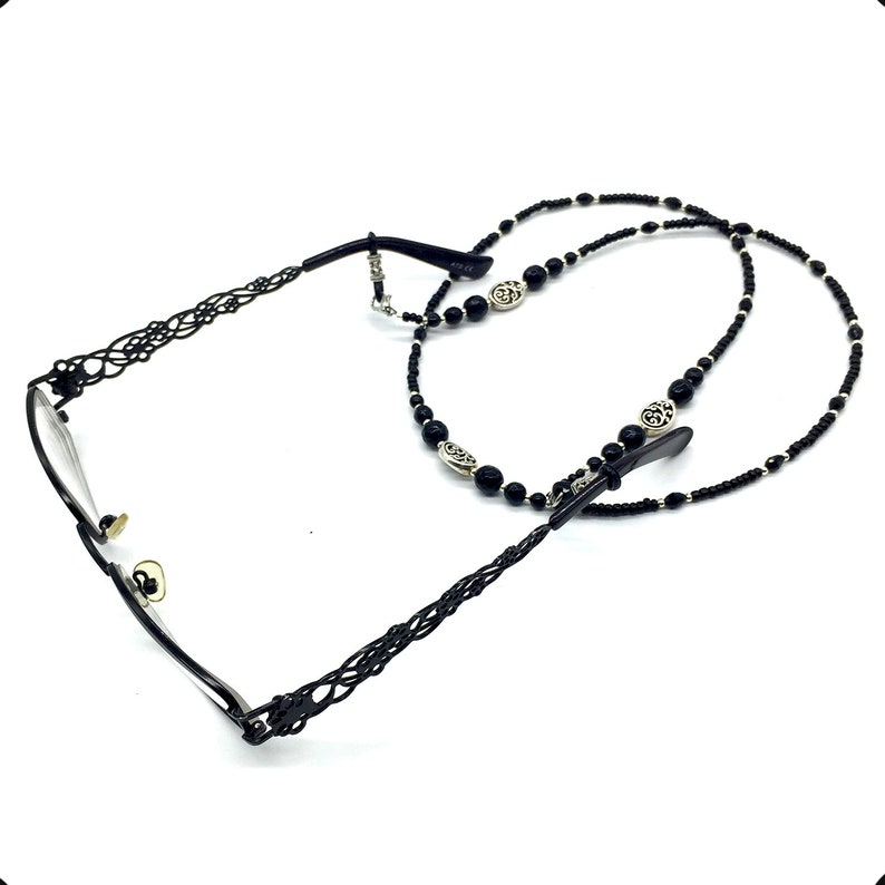 May include: Black beaded eyeglass chain with silver-toned charms. The chain is made of small black beads and features a decorative metal design. The chain is attached to the glasses with two small metal clips.