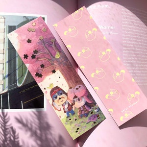 May include: Two illustrated bookmarks. One features a colorful scene with cartoon characters under a cherry blossom tree. The other is pink with a repeating yellow cartoon character pattern. The text "shivi studio" is visible on one bookmark.