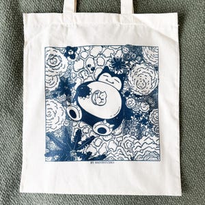 May include: White tote bag featuring a blue and white illustration of a cartoon character with a smaller character inside. The design is surrounded by floral and abstract elements. The text "BY SHIYISTUDIO" is at the bottom of the design.