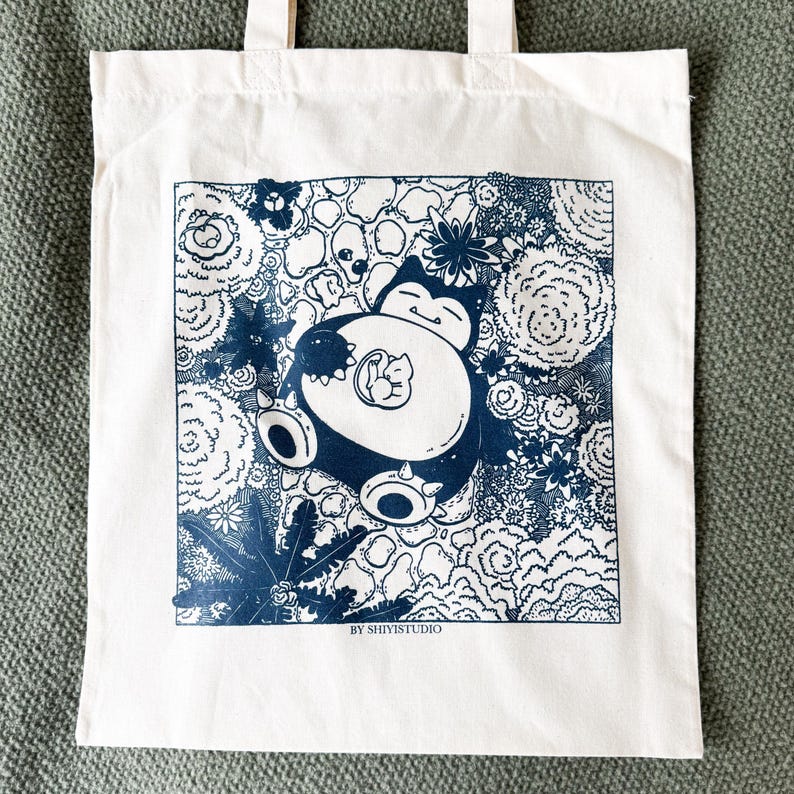 May include: White tote bag featuring a blue and white illustration of a cartoon character with a smaller character inside. The design is surrounded by floral and abstract elements. The text "BY SHIYISTUDIO" is at the bottom of the design.