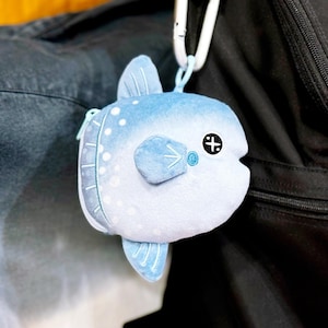 以下が含まれることがあります： 青い色合いに白い水玉模様が施された、マンボウ型のぬいぐるみコインケース。小さな横びれ、背びれ、黒と白の十字型の目が特徴です。柔らかく丸みを帯びた魚の形をしたポーチには、取り付け用の白いプラスチック製クリップが付いています。