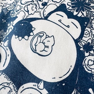 May include: A white fabric with a navy blue illustration of a sleeping Snorlax with a Mew inside. The design is surrounded by floral and other cartoon elements. The image is a close-up of the fabric.