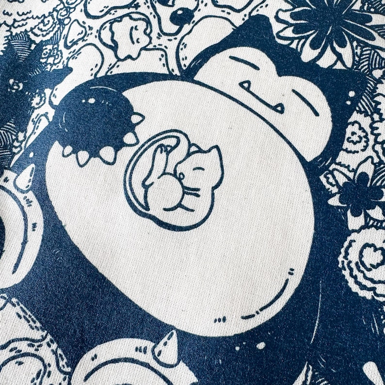 May include: A white fabric with a navy blue illustration of a sleeping Snorlax with a Mew inside. The design is surrounded by floral and other cartoon elements. The image is a close-up of the fabric.