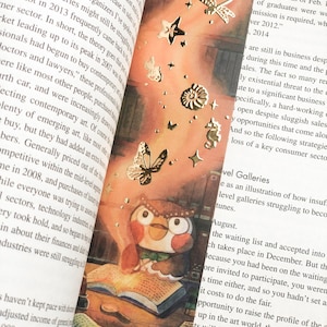 Puede incluir: Una ilustraci&oacute;n de acuarela de un b&uacute;ho marr&oacute;n con mejillas rosadas leyendo un libro. El b&uacute;ho est&aacute; rodeado de libros y otros objetos, y hay estrellas doradas y mariposas esparcidas alrededor de la imagen. El texto "Lawyers" y "Nationally, 11.2 percent of [law school] graduates were working as of Feb. 2014." es visible en el fondo.