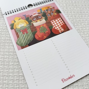 K&ouml;nnte beinhalten: Ein Spiral-Kalender mit einer Dezemberseite mit Illustrationen von Cartoon-Charakteren in Weihnachtsstr&uuml;mpfen. Die Str&uuml;mpfe sind gr&uuml;n, rot und wei&szlig;, vor einem rosa Hintergrund. Das Wort "Dezember" ist in roter Schrift geschrieben.