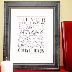 Peut inclure: Cadre avec l'inscription "NEVER STOP PRAYING, BE THANKFUL..." en calligraphie. Une bougie blanche et un grand vase rouge sont posés sur une surface rouge, avec un mur jaune clair en arrière-plan.