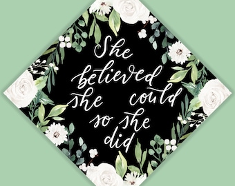Può includere: Decorazione per cappello di laurea con la scritta "She believed she could so she did" in corsivo bianco su sfondo nero. Ornato con rose bianche e foglie verdi, su sfondo verde chiaro.