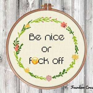 May include: A white cross stitch embroidery hoop with a floral wreath design. The text "Be nice or f*ck off" is stitched in black thread with a yellow smiley face emoji.