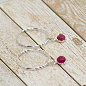 May include: A pair of silver hoop earrings with a small, round, red gemstone dangling from each hoop.
