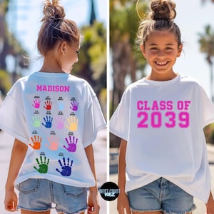 May include: A white t-shirt featuring a pink graphic on the front that reads CLASS OF 2039. The back displays the name MADISON in pink above twelve colourful handprint graphics, each labelled with a grade level from kindergarten to 12th grade.