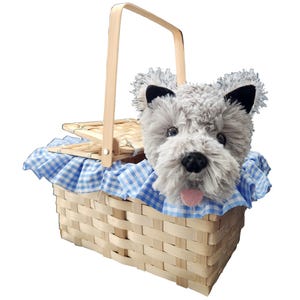 May include: A light-colored woven wooden basket with a swing handle and a blue and white gingham fabric liner. Inside the basket sits a plush, grey, shaggy-textured dog figure with black ears, a black nose, and a small pink tongue sticking out.