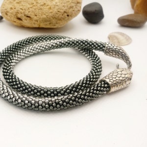 May include: A silver and black beaded necklace with a snake-shaped clasp. The necklace is made of small, round beads that are arranged in a graduated pattern, with the silver beads at the top and the black beads at the bottom.