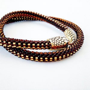 Op de afbeelding: Een bruine en gouden kralenketting met een decoratieve metalen sluiting. De ketting is gemaakt van kleine, ronde kralen die in een patroon zijn geweven.