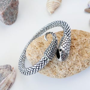 Puede incluir: Un brazalete de serpiente plateado con un dije de cabeza de serpiente detallado. El brazalete está hecho de pequeñas cuentas plateadas y está enrollado alrededor de una roca marrón claro.