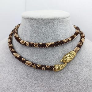 May include: A beaded necklace in shades of brown and beige, displayed on a mannequin. The necklace has a snake-shaped clasp in gold. The beads are tightly woven, creating a textured pattern. The necklace is designed to be worn around the neck.