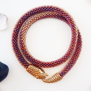 Peut inclure: Un collier de perles avec un fermoir en forme de tête de serpent. Le collier est composé de deux rangs de perles, l'un brun et l'autre violet. Le fermoir en forme de tête de serpent est de couleur dorée.