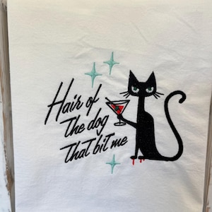 May include: White hand towel with a black cat illustration holding a martini glass. The text "Hair of the dog that bit me" is embroidered in black, with teal star accents and red blood drops.