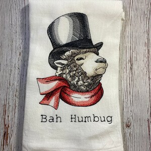 May include: White kitchen towel with an embroidered image of a sheep wearing a top hat and a red scarf. The text "Bah Humbug" is embroidered below the image.