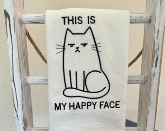 May include: White kitchen towel with black embroidery of a cat with the text "This is my happy face".
