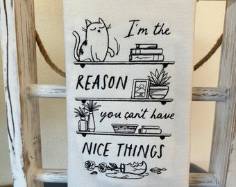 May include: A white kitchen towel with black text and illustrations. The text reads "I'm the reason you can't have nice things". The illustration is of a cat knocking over a shelf with plants, books, and a picture frame.