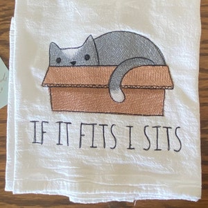 May include: White kitchen towel with an embroidered gray cat sitting in a brown box. The text "If it fits I sits" is embroidered below the cat.