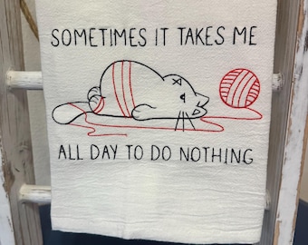 May include: White kitchen towel with black embroidery that reads "Sometimes it takes me all day to do nothing". A cartoon cat is embroidered in black with red stripes and a ball of yarn in red and white.