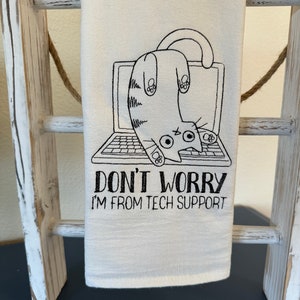 May include: A white tea towel with a black embroidered design of a cat hanging upside down from a laptop with the text "Don't Worry I'm From Tech Support".