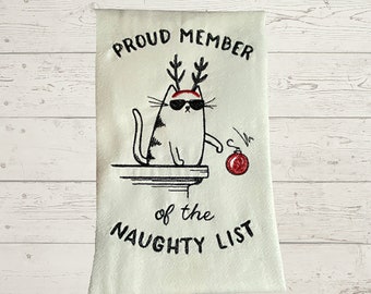 May include: White kitchen towel with black embroidery of a cat wearing sunglasses and reindeer antlers. The text reads "Proud Member of the Naughty List".