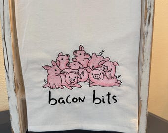 May include: White kitchen towel with pink embroidered pigs and the text "bacon bits".