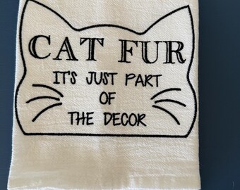 May include: White folded kitchen towel with black embroidered text and a cat face outline. The text reads "CAT FUR IT'S JUST PART OF THE DECOR". The towel is made of a textured fabric.