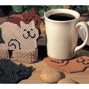 Könnte beinhalten: Vier gehäkelte Untersetzer in Katzenform, zwei in Beige, einer in Braun und einer in Schwarz. Die Untersetzer liegen auf einer Holzoberfläche neben einer weißen Tasse mit Kaffee und einem Teller mit Keksen.