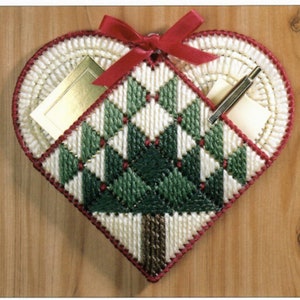 Puede incluir: Un lienzo blanco en forma de corazón con un patrón de cuadros verde y blanco, un lazo rojo y dos bolsillos. Los bolsillos están llenos de una tarjeta dorada y un bolígrafo.