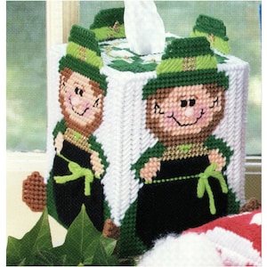 May include: A green and white crocheted tissue box cover featuring two smiling leprechauns wearing green hats with gold trim. The leprechauns are holding pots of gold.