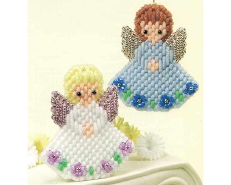 May include: Two handmade plastic canvas angel ornaments. One angel is white with pink wings and a light pink heart. The other angel is light blue with silver wings and a light pink heart. Both angels have a floral border with purple and blue flowers.