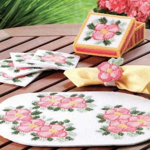 May include: A set of pink and green floral-themed plastic canvas coasters, a matching napkin ring, and a tissue box cover. The items are all on a wooden table.