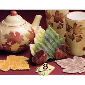 May include: A set of four crocheted leaf-shaped coasters with acorn accents. The coasters are in shades of green, brown, yellow, and beige. The coasters are displayed on a red surface with a teapot and a mug in the background.