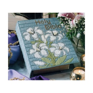 May include: A blue and white canvas bible cover with a floral design of white lilies and green leaves. The cover reads "Holy Bible".