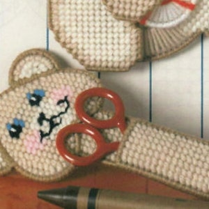 May include: A beige crocheted teddy bear with blue eyes and a pink nose holding a pair of red plastic scissors.