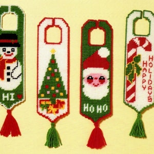 May include: Four Christmas-themed door hangers with different designs. The first features a snowman with the word "HI". The second features a Christmas tree with presents. The third features Santa Claus with the words "HO HO HO". The fourth features a candy cane with holly and the words "HAPPY HOLIDAYS".