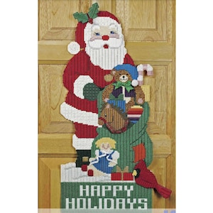 May include: A plastic canvas Christmas decoration featuring Santa Claus carrying a bag of toys. The bag is filled with a teddy bear, a rocking horse, a doll, and a cardinal. The decoration also includes the text "HAPPY HOLIDAYS".