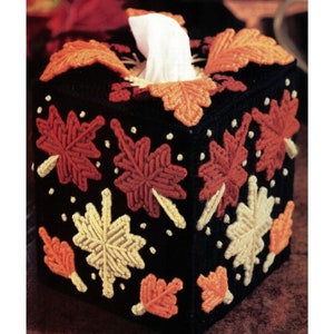 May include: A black tissue box cover with a fall leaf design. The leaves are made of yarn in shades of orange, red, and yellow. The cover is decorated with small white dots.