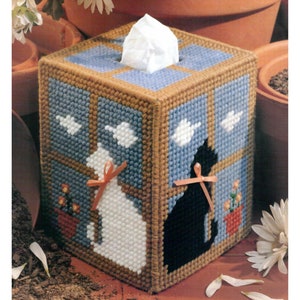 May include: A blue and brown tissue box cover with a window scene featuring two cats, one white and one black, looking out the window. There are white birds flying in the sky and flowers in pots on the windowsill.