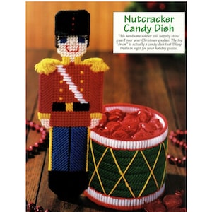 May include: A red and white plastic nutcracker soldier holding a green and white candy dish shaped like a drum. The candy dish is filled with red candies. The text "Nutcracker Candy Dish" is printed above the image.