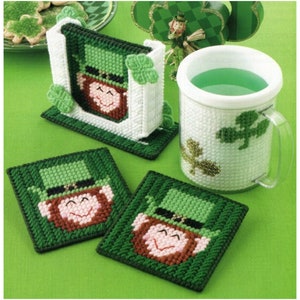 May include: A St. Patrick's Day themed set including a mug, coasters, and a napkin holder. The coasters and napkin holder feature a leprechaun face and are green, white, and brown. The mug is white with green shamrock accents. The items are on a green surface.