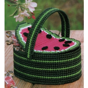 May include: A green and red crocheted watermelon-shaped basket with a handle. The basket is filled with wrapped candy.